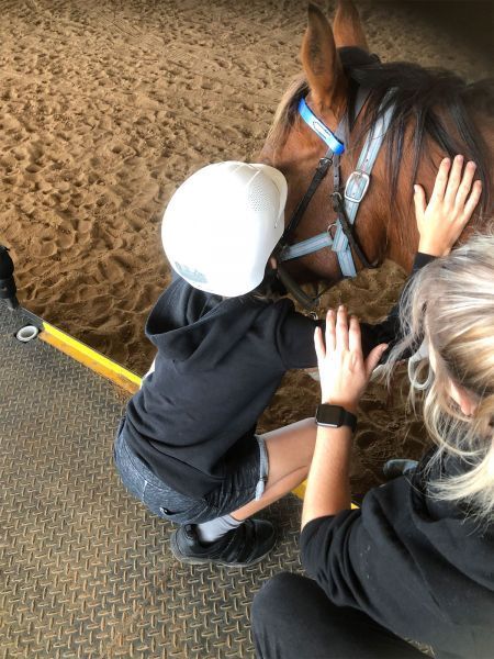 Child in helmet and adult petting a brown horse in a sandy indoor arena.