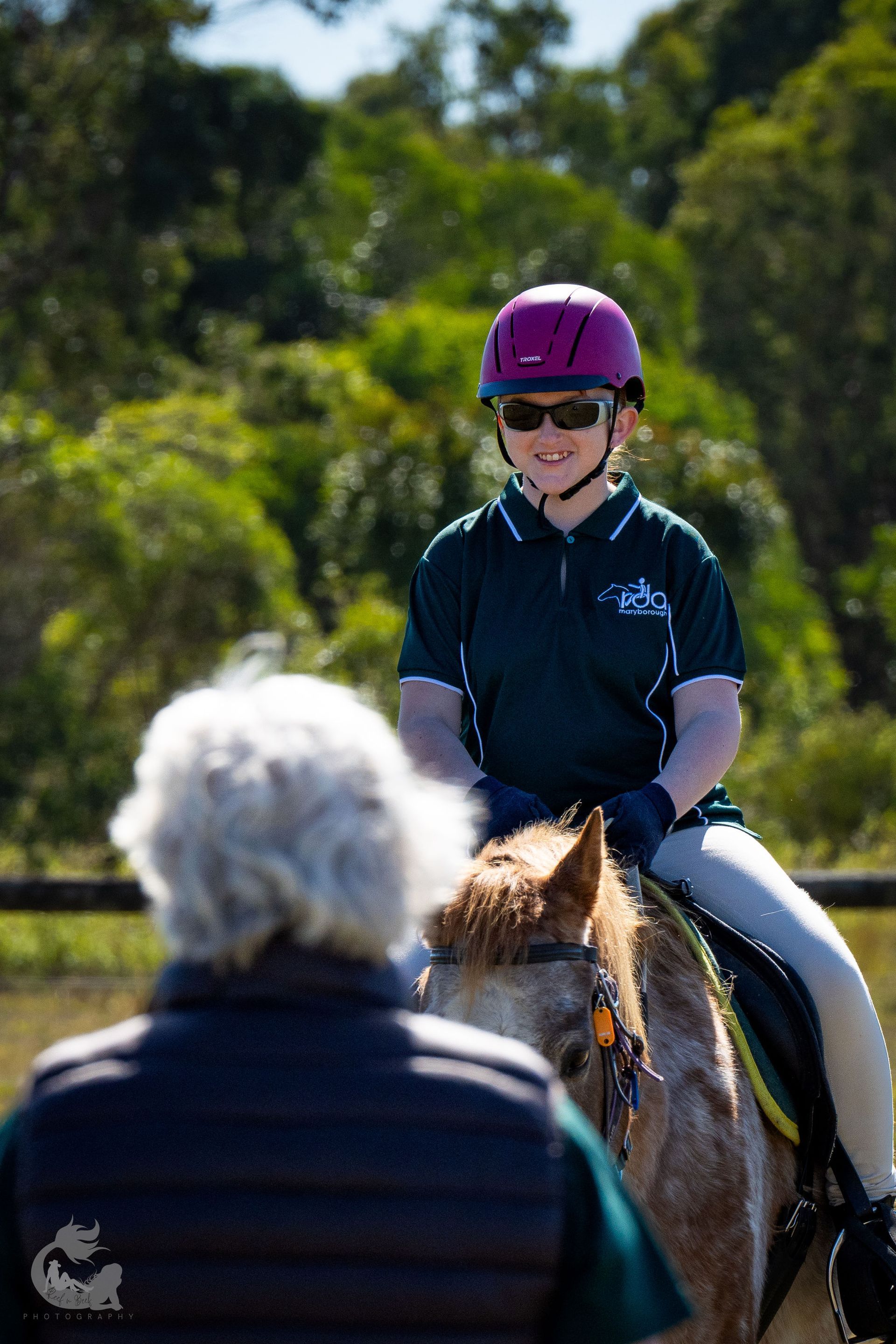 Young rider on horse wearing helmet, being observed by instructor in outdoor setting.