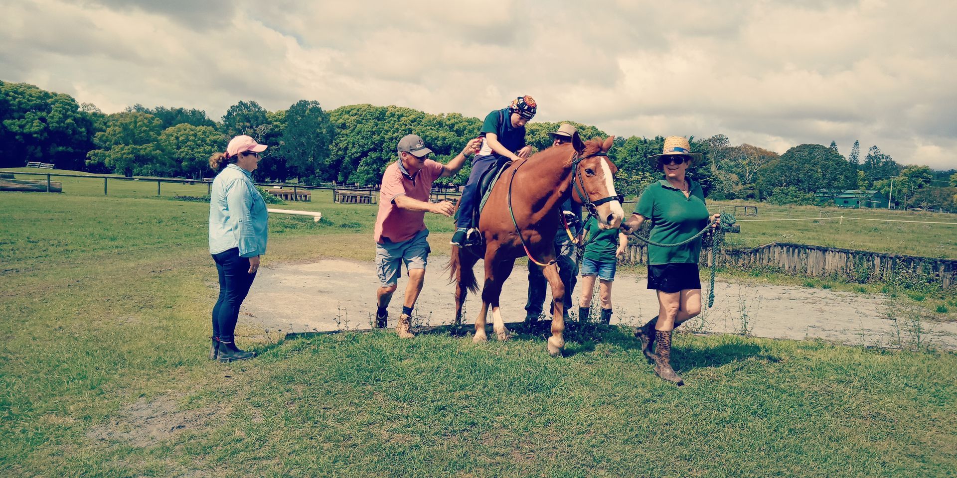 People helping a rider on a horse. They stand on green grass under a cloudy sky.