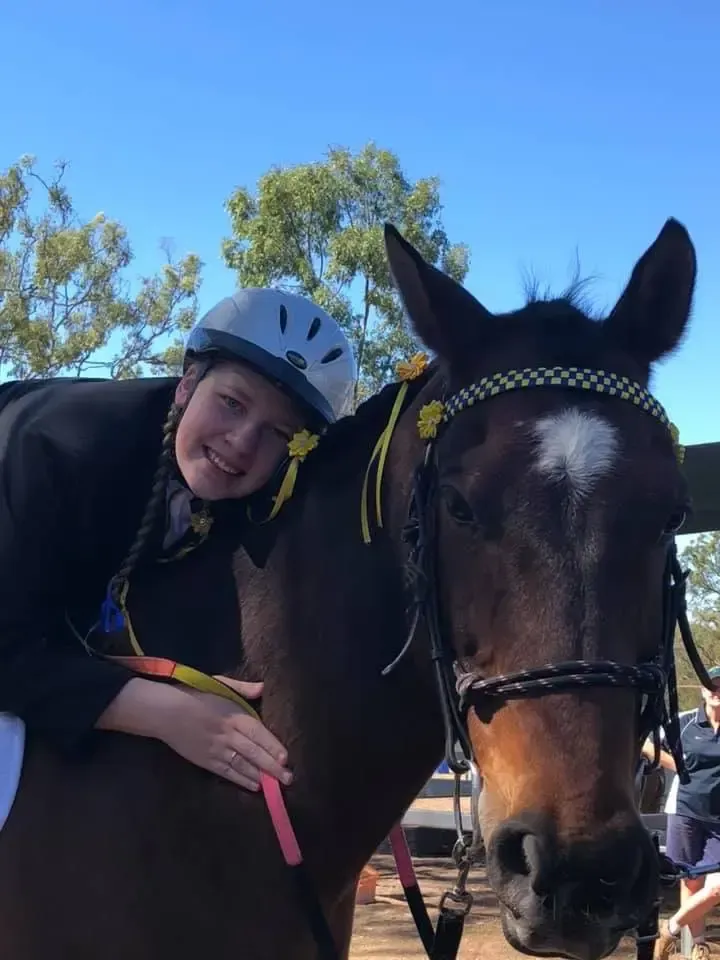 A person in a helmet hugs a dark horse with yellow ribbons; both smile under a blue sky.
