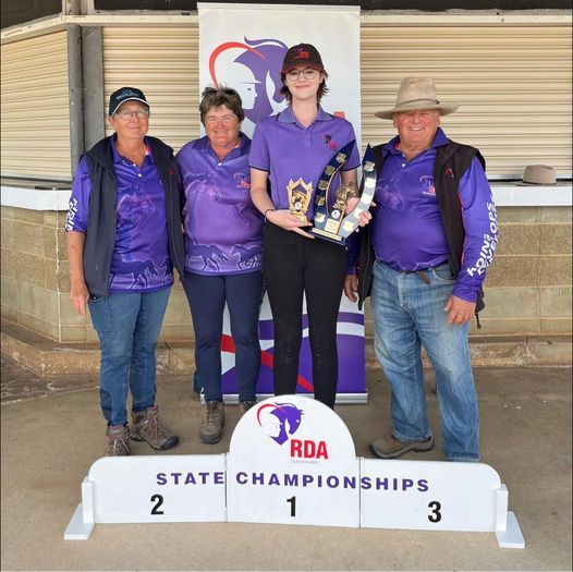 People with trophies on a podium: RDA State Championships. The person in the middle holds trophies. Others wear purple shirts.