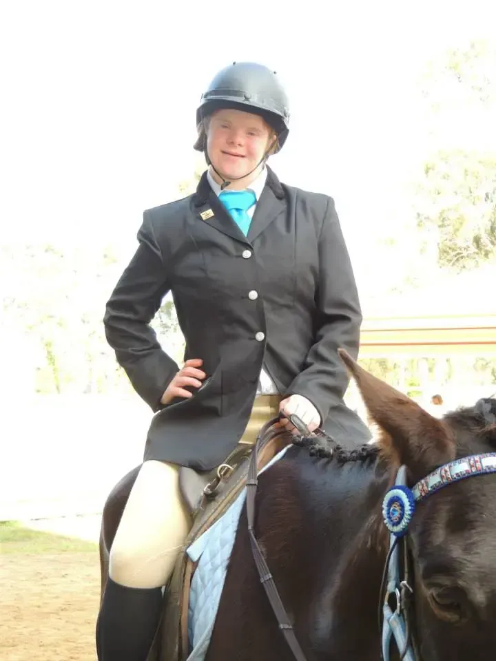 Person on horseback in riding attire, smiling. Outdoor setting. Horse has ribbon on bridle.