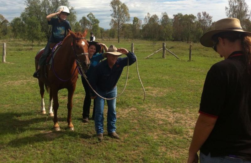 Person on horse, others assisting with gear in a grassy field; blue skies and trees in the background.