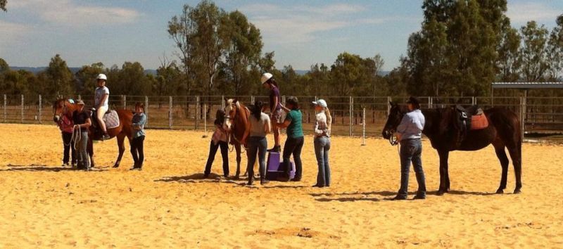People and horses in a riding arena, some are helping riders mount the horses.