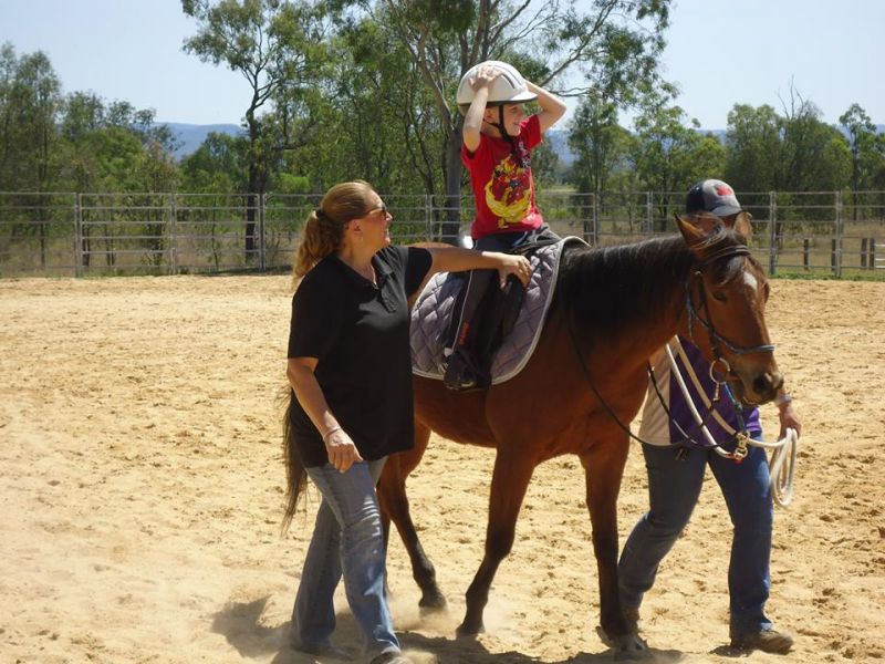Child on horseback with two adults assisting in a sandy riding area.