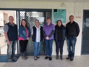 Six people standing in front of a building with a plaque. They are casually dressed and smiling.