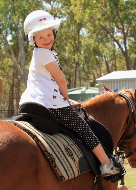 Girl in helmet smiles while riding a brown horse outdoors.