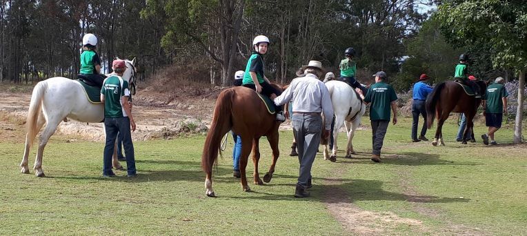 Children riding horses, guided by adults on a grassy field; trees in the background.