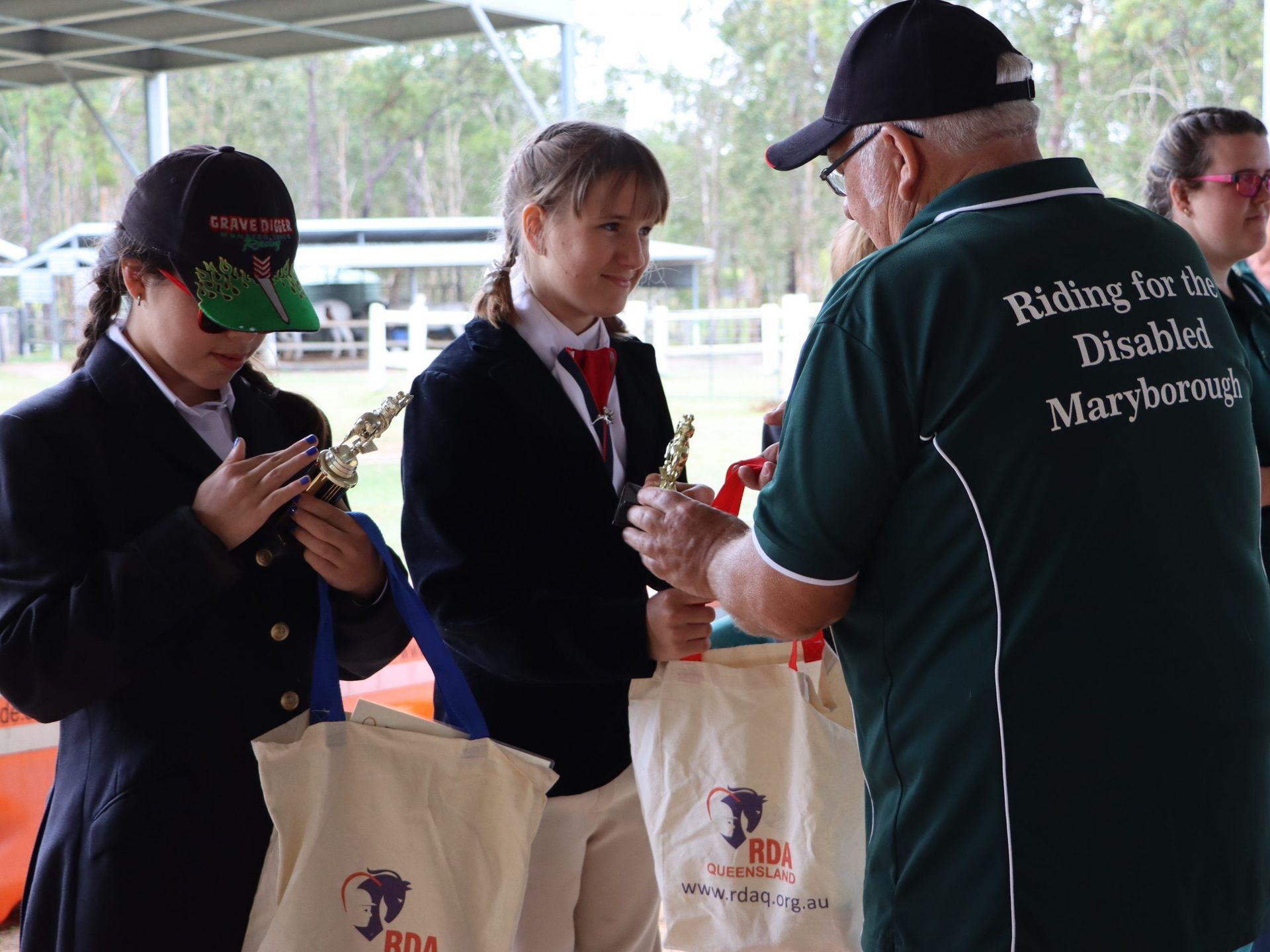 Two people receive trophies from a man at an RDA event. They wear riding attire and hold gift bags.