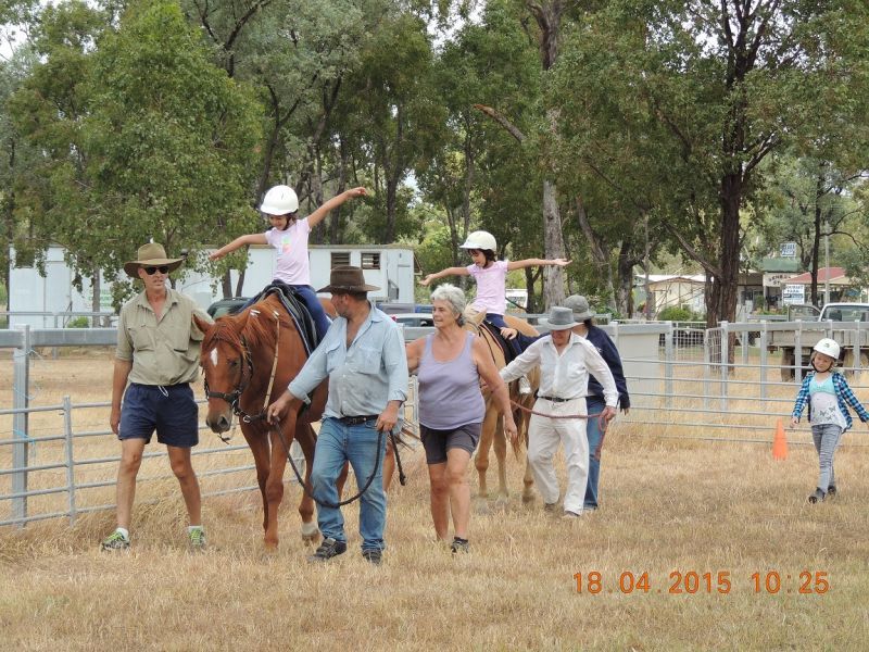 People leading two children on horseback, arms outstretched, in a grassy area near a fence.