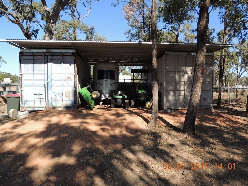 Three cargo containers shelter machinery, under a corrugated metal roof, trees in the background, blue sky.