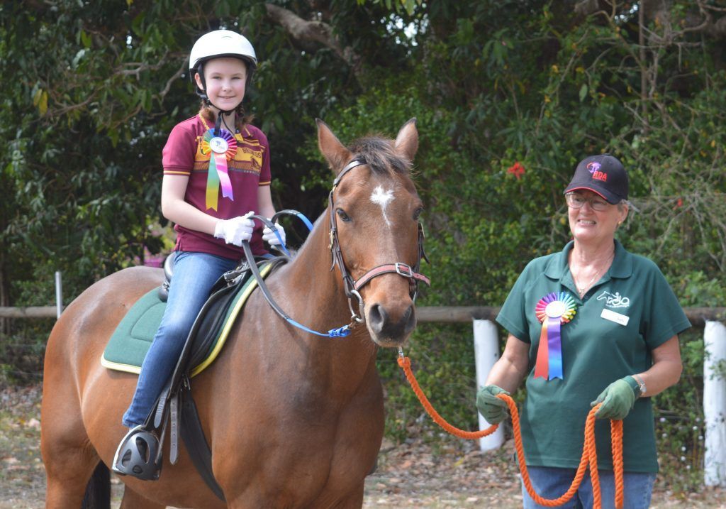 Girl riding a brown horse guided by a woman; both wearing ribbons in an outdoor setting.