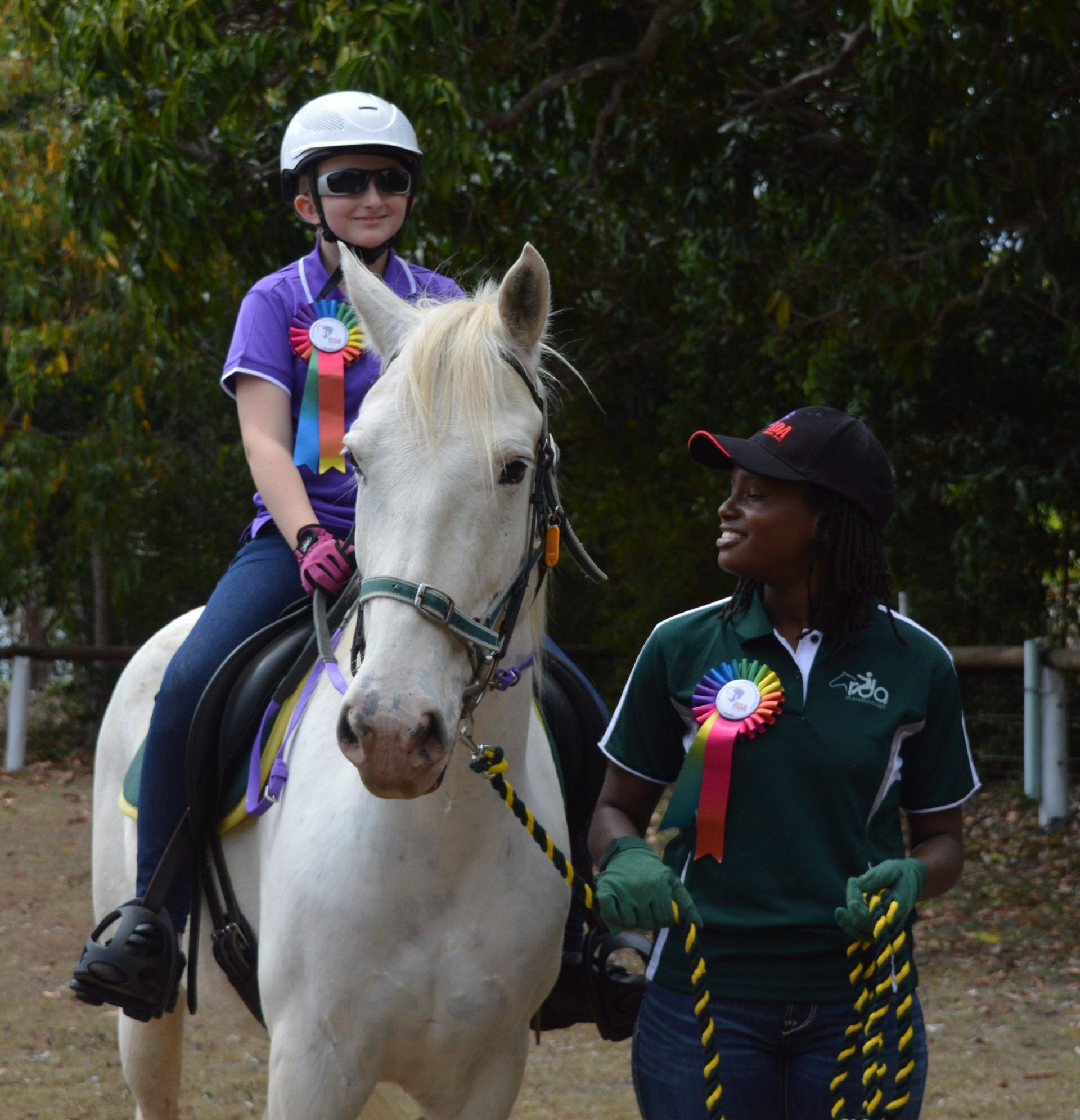 Young person on white horse, being led by a person with a ribbon, both smiling, outdoors.