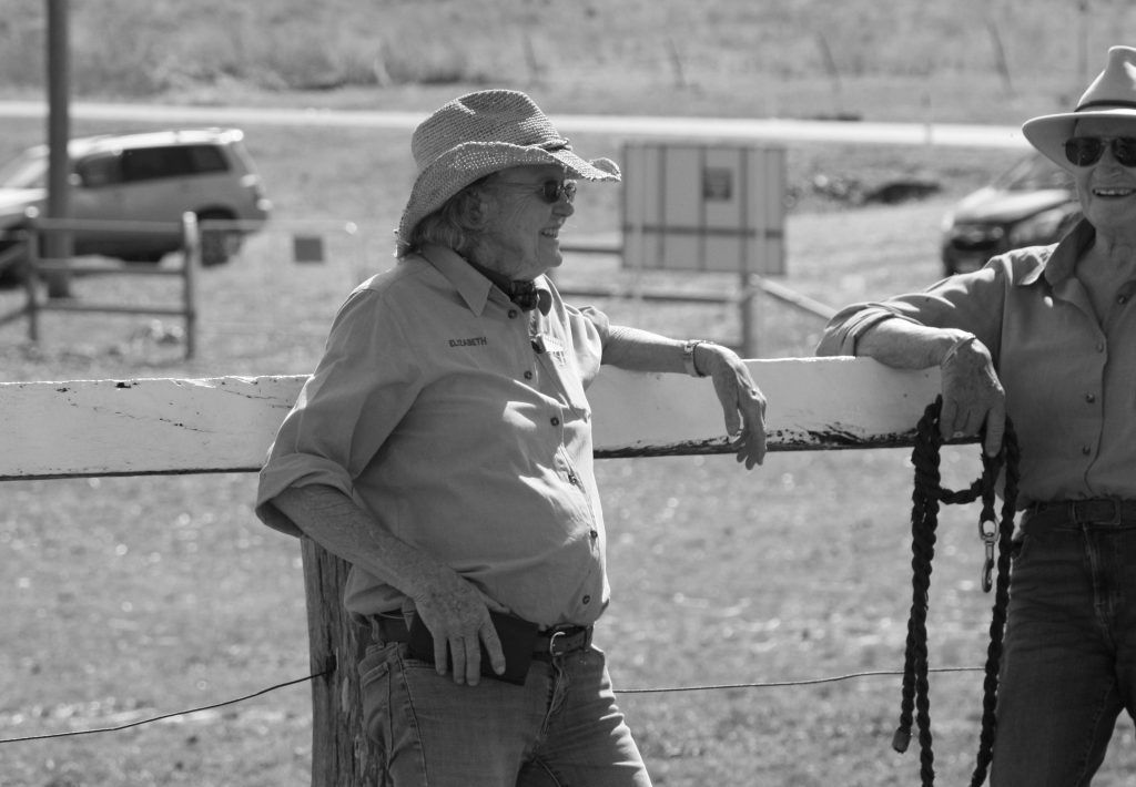 Two people in cowboy hats lean on a fence, outdoors.