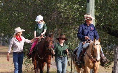 People riding horses on a trail, accompanied by a person leading a horse, sunny day.