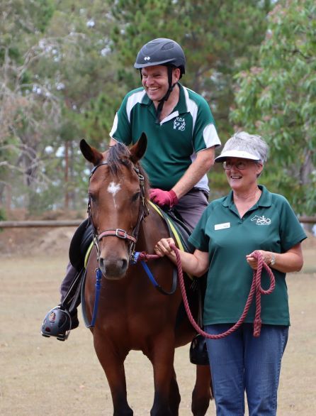 Man on brown horse smiles as woman leads, both wearing green shirts, outdoors.