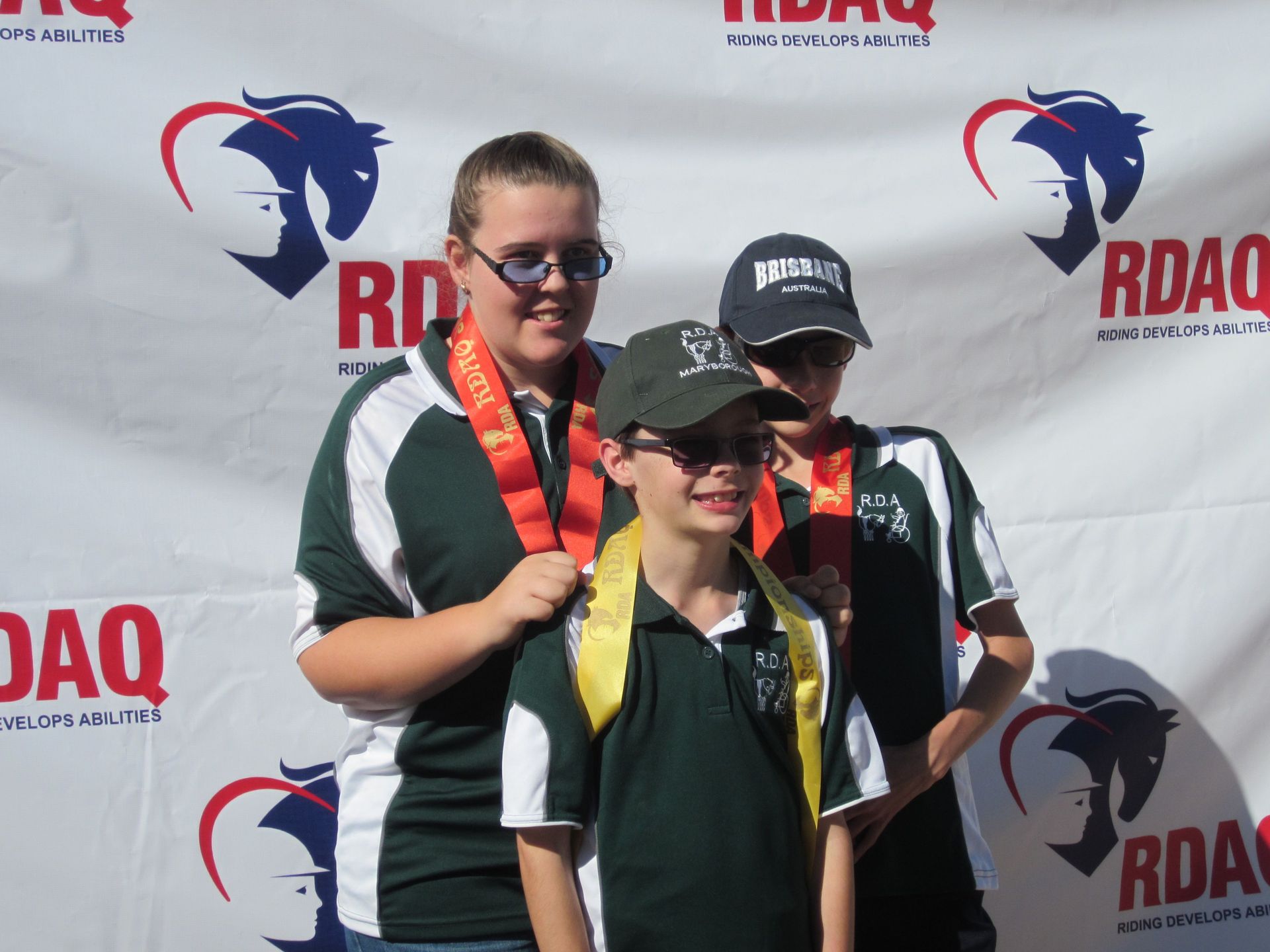 Three people wearing medals pose in front of an RDAQ backdrop. Green and white shirts. Sunglasses.