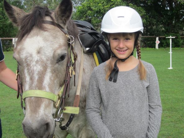 Girl in riding helmet smiles next to a horse with saddle in an outdoor setting.