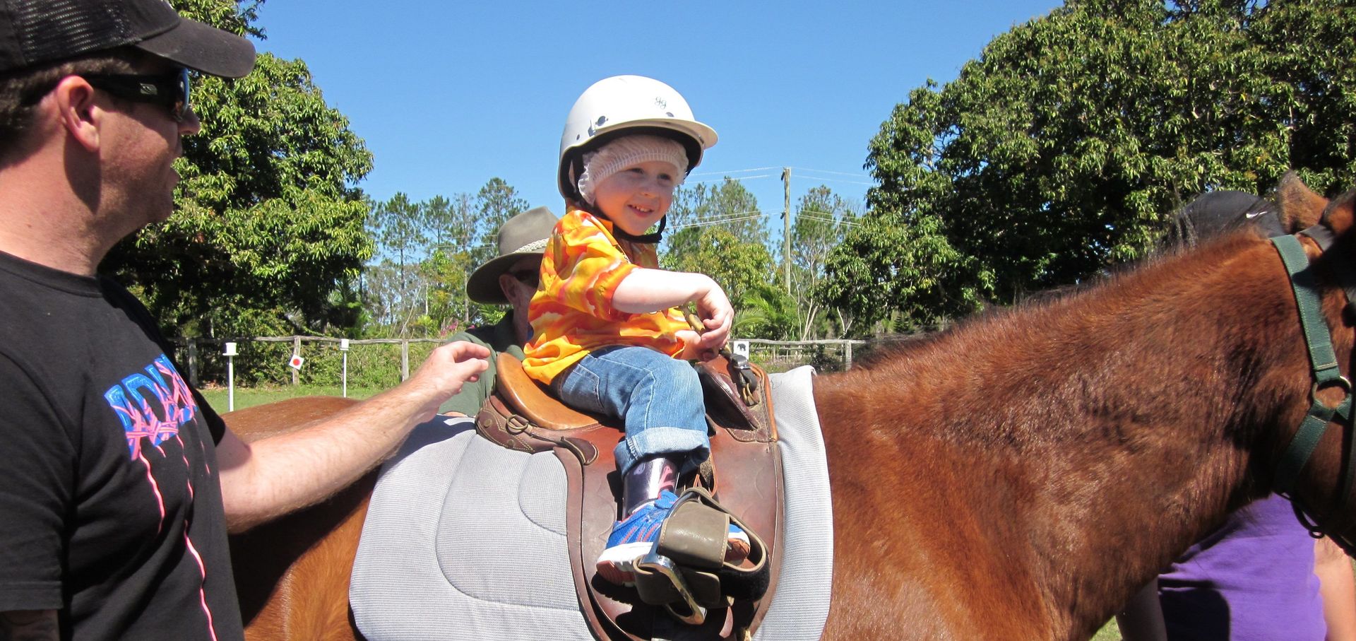 A child in a white helmet and orange shirt sits on a horse with a man next to them.