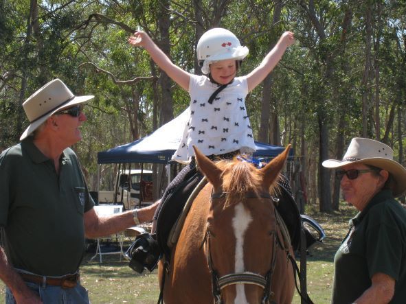 Girl on a horse with arms raised, helmet on, cheered on by two adults in a sunny outdoor setting.