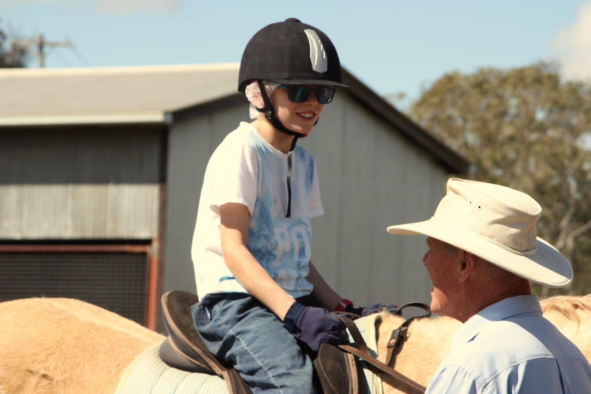 Boy on horse, wearing helmet and sunglasses, talking to person in a hat, outdoors near a building.