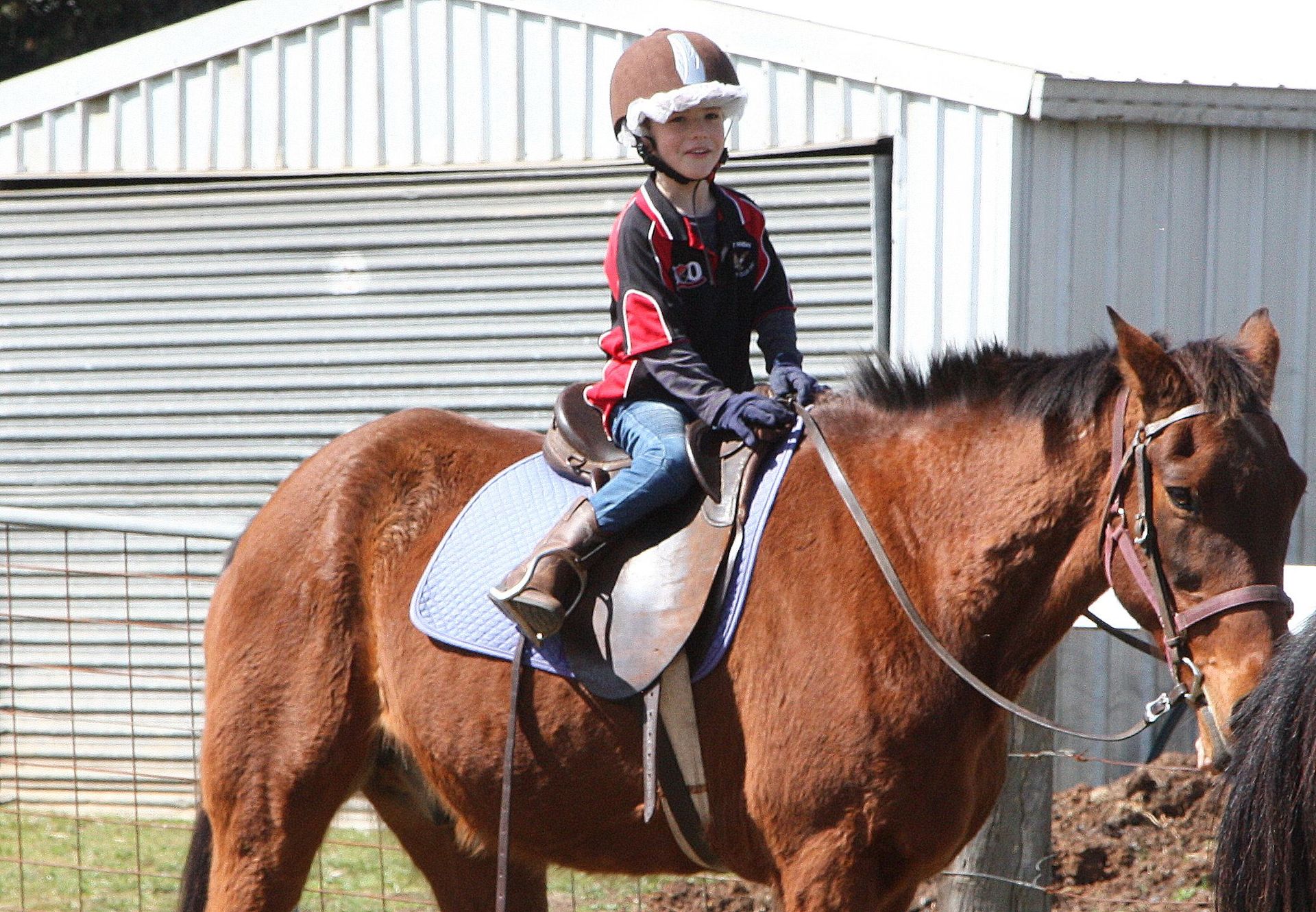 Young person on a brown horse, wearing helmet and boots, outside a shed.