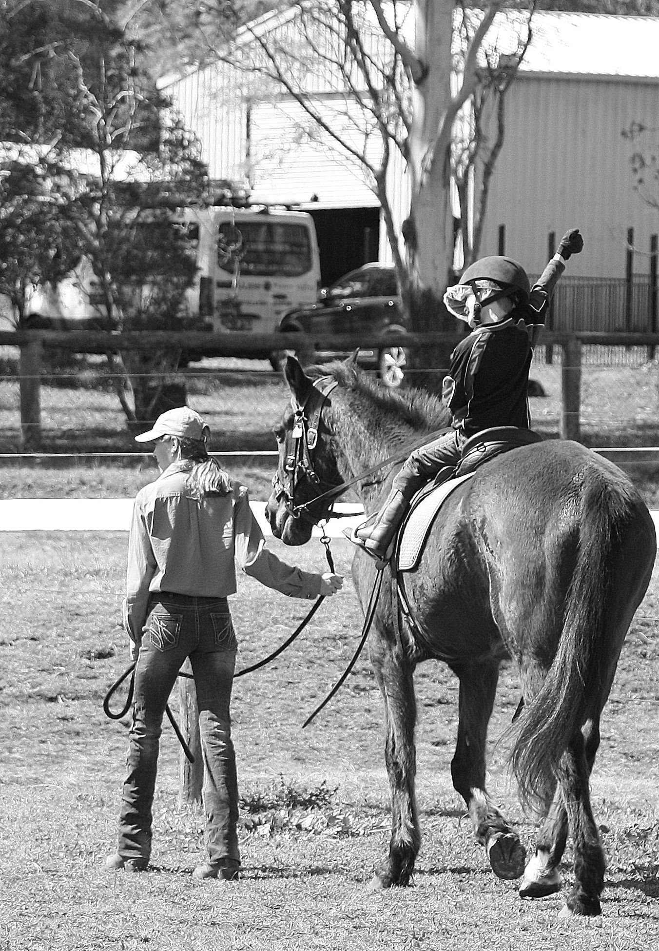A child on horseback with a handler, waving a fist. Horse is brown, and setting is outdoors.