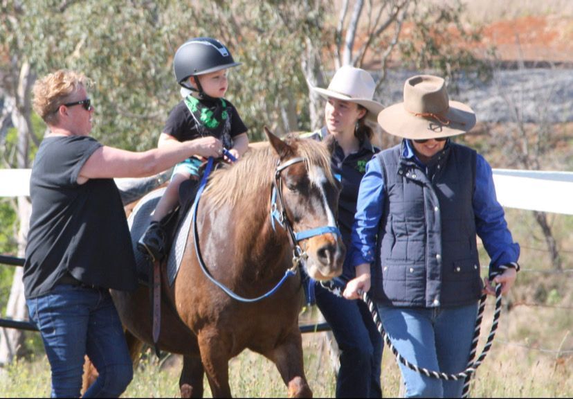 Child on horseback, supported by three adults, outdoors.