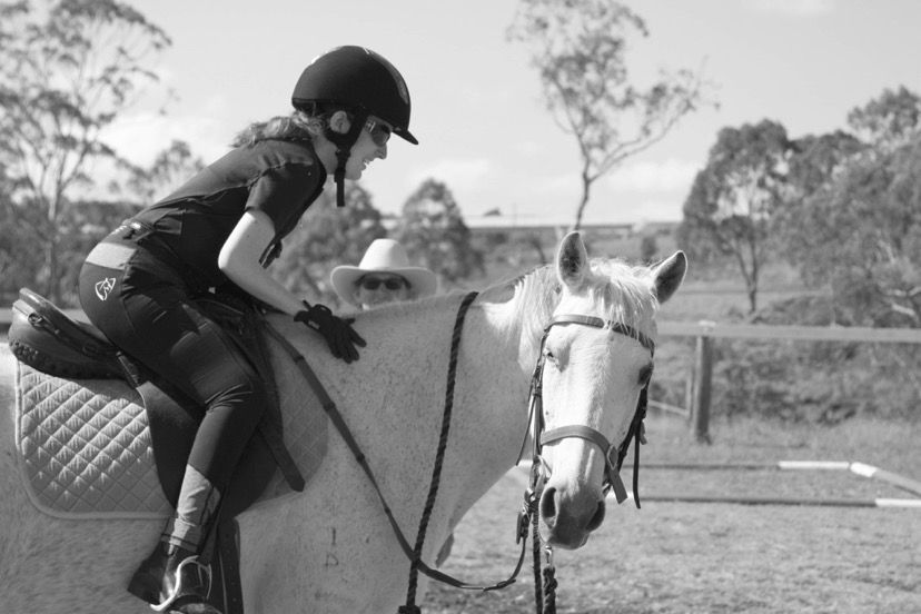 Person riding a white horse in an outdoor arena, wearing a helmet and gloves.