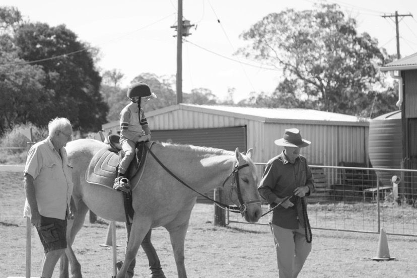 A child rides a horse with a helmet, guided by two adults in an outdoor setting.
