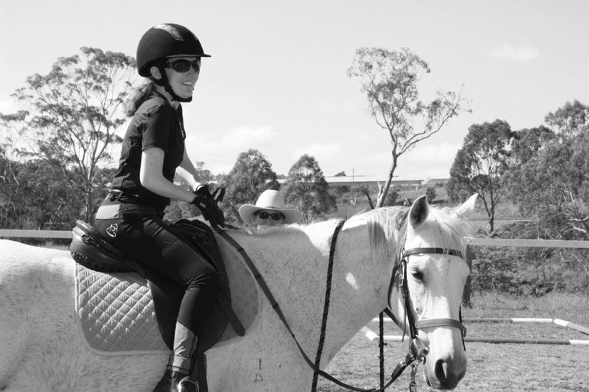 Woman riding a white horse outdoors, wearing a helmet and sunglasses.