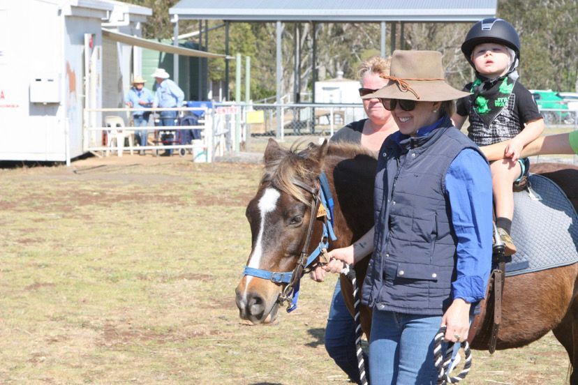 Woman leading a pony with a child wearing a helmet on its back at an outdoor event.