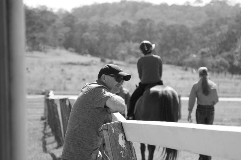 Man leaning on fence watches person riding horse; another person walks nearby in rural setting.