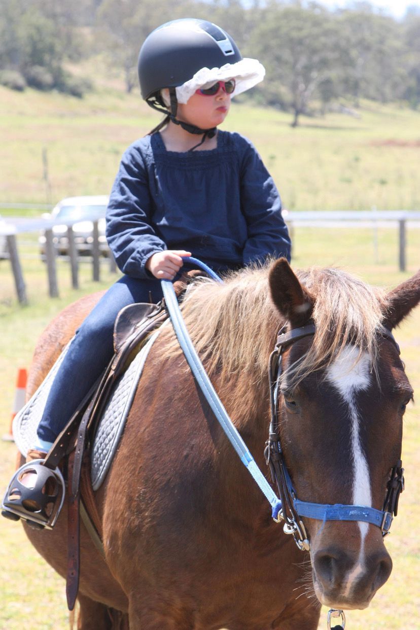 Child riding a brown pony, wearing a helmet and sunglasses, outdoors on a sunny day.