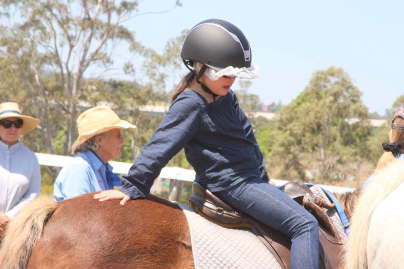 Child wearing helmet on a pony, assisted by an adult. Outdoors, sunny.