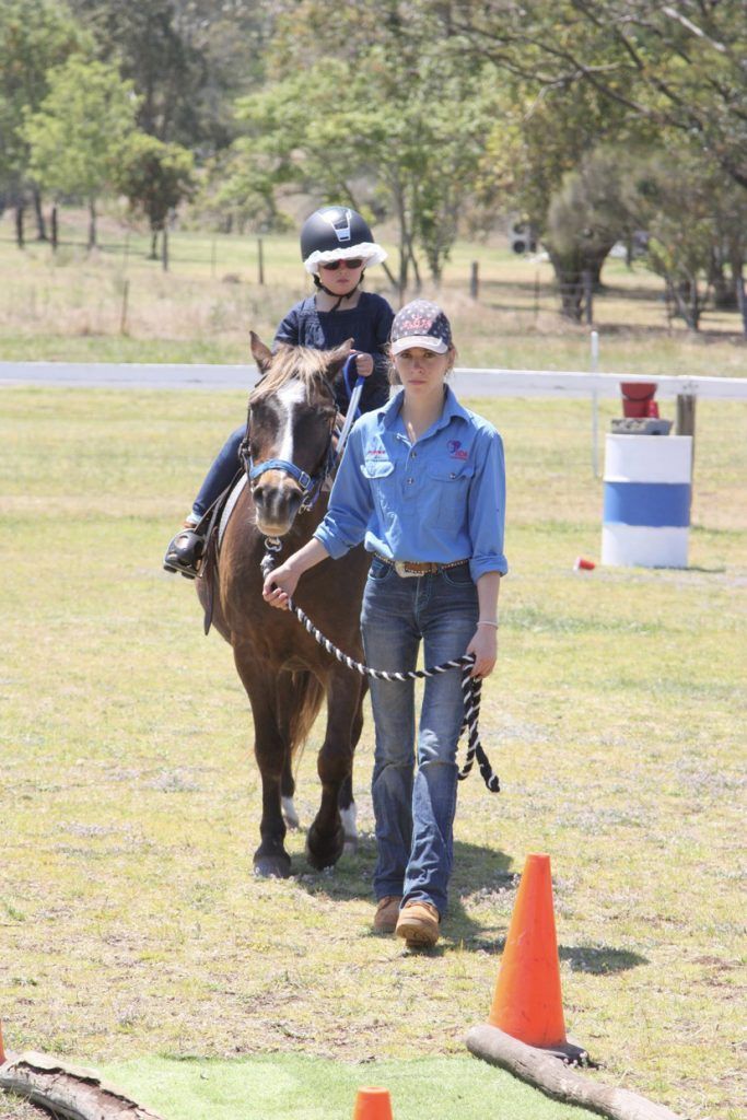 Young child riding a pony, led by a person in a blue shirt and jeans on a grassy field.