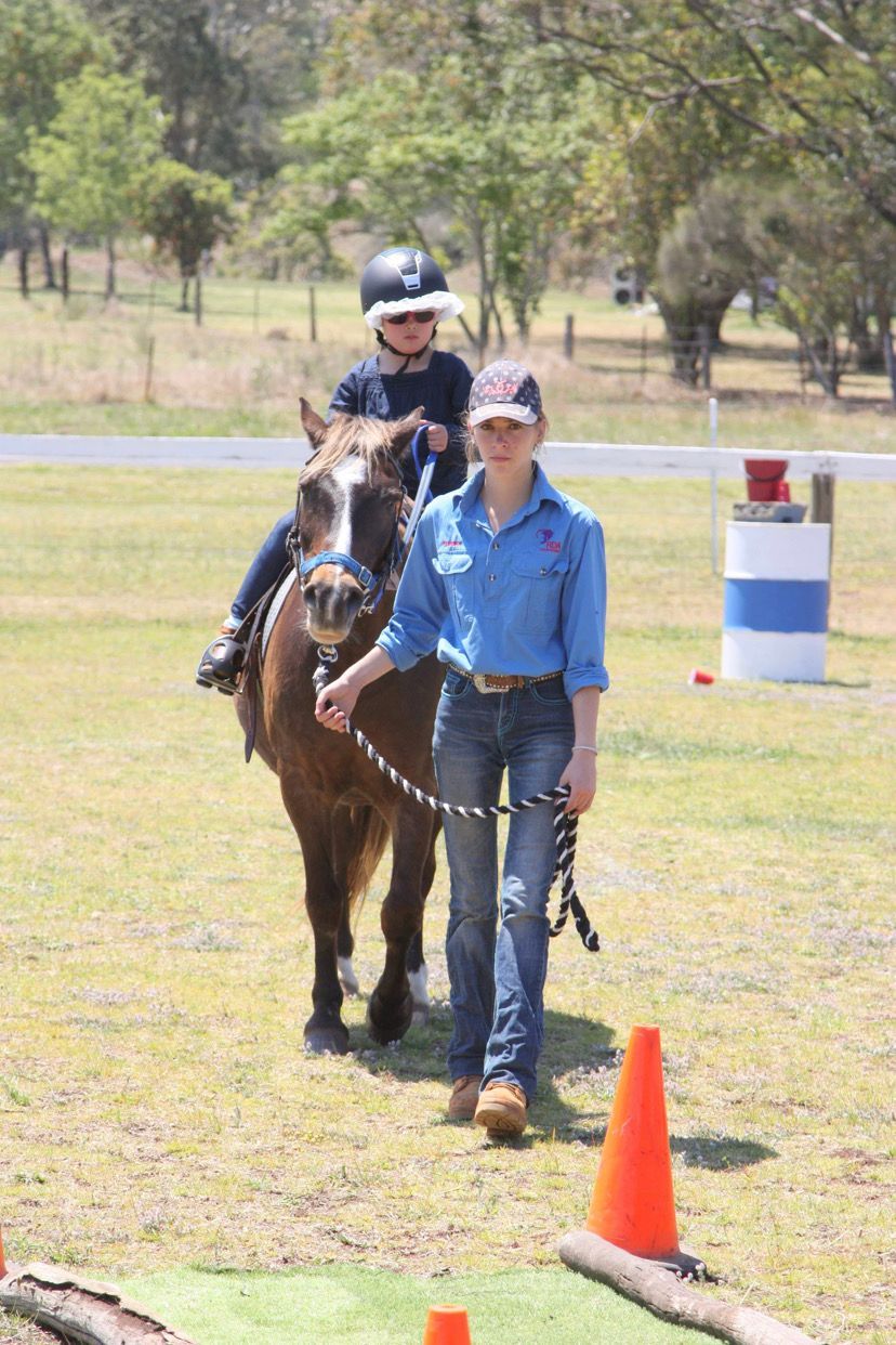 A young person rides a brown horse, led by a person in a blue shirt and jeans on a grassy field.