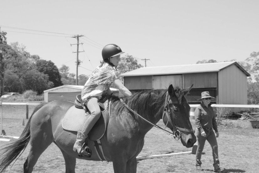Young person riding a brown horse in an outdoor arena, overseen by an instructor.