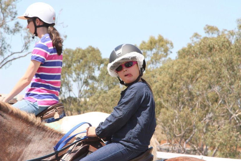 Two people horseback riding; girl looks at camera with trees and blue sky background.