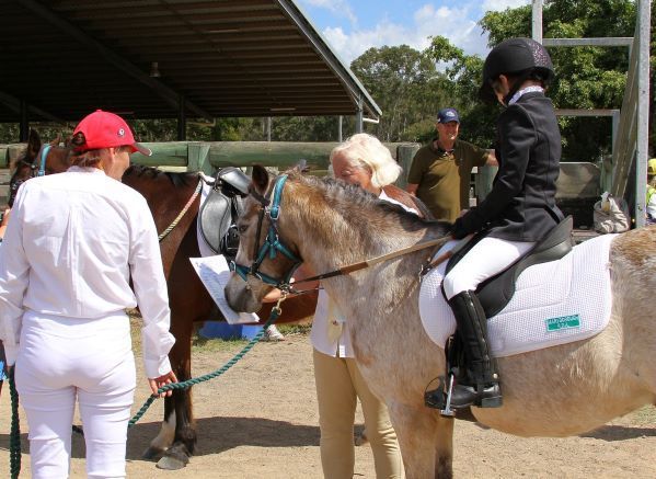 Child on a pony, with instructor adjusting its bridle. Others look on, in an outdoor equestrian setting.