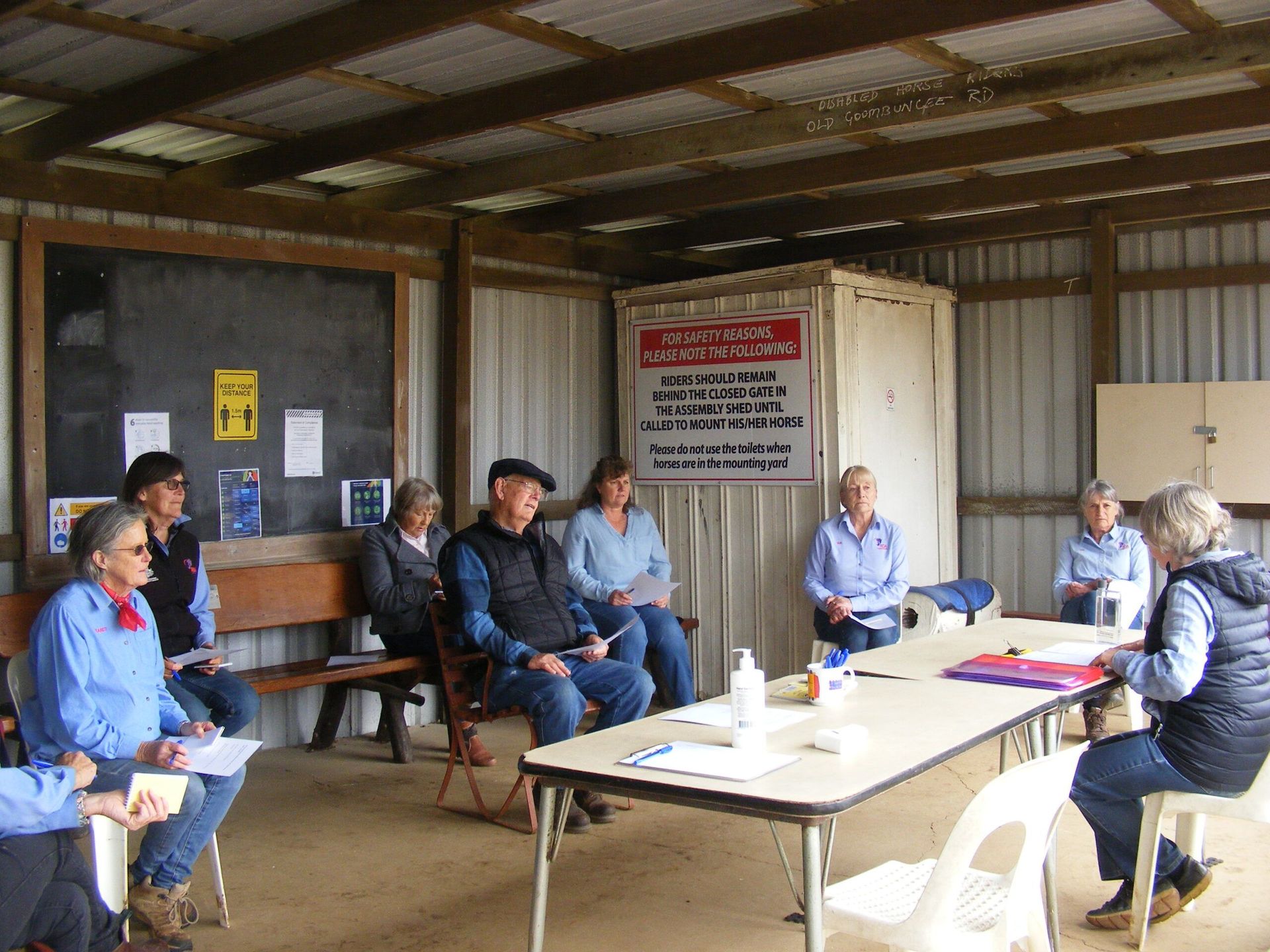 People seated around a table in an open shed, possibly a meeting or workshop.