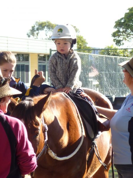 Child wearing a helmet sits on a brown horse, being assisted by adults. Outdoors.