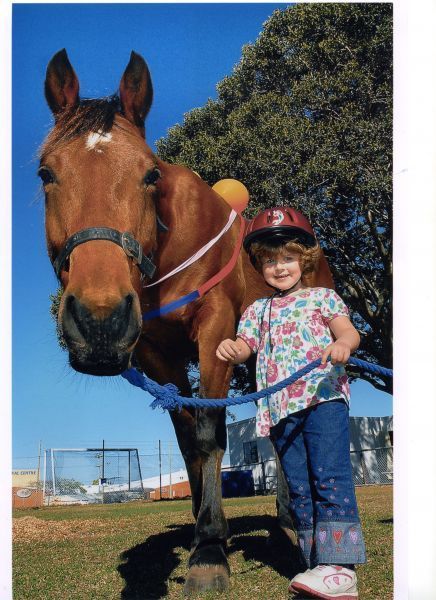 Girl in helmet smiles next to brown horse, holding a blue lead rope, outdoors on a sunny day.