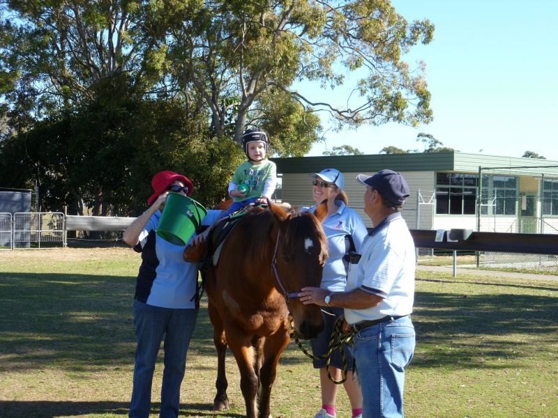 Child riding a brown horse; adults assist, pouring water. Outdoors, sunny.