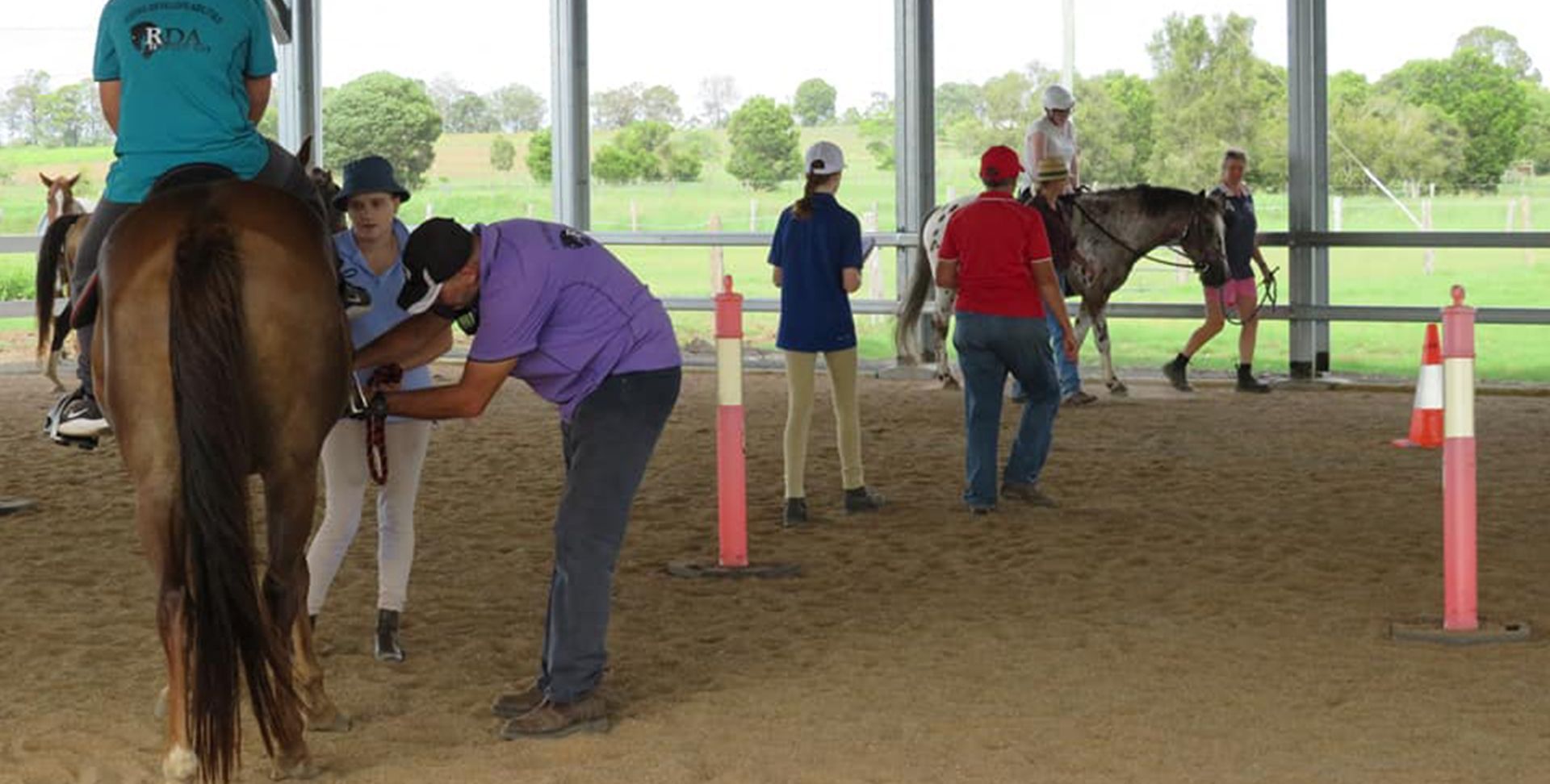 People in an arena with horses. A man adjusts tack, others ride or watch.