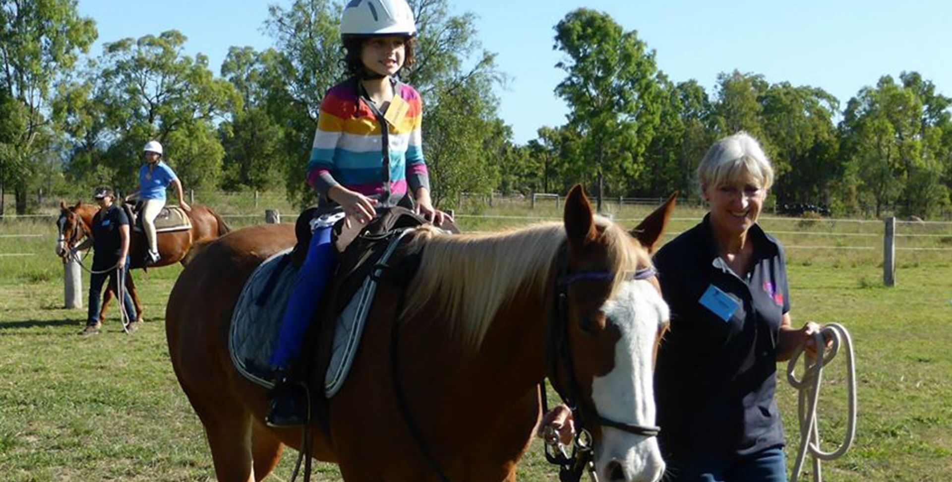 Girl on a horse led by a woman, both smiling, outdoors on a sunny day.