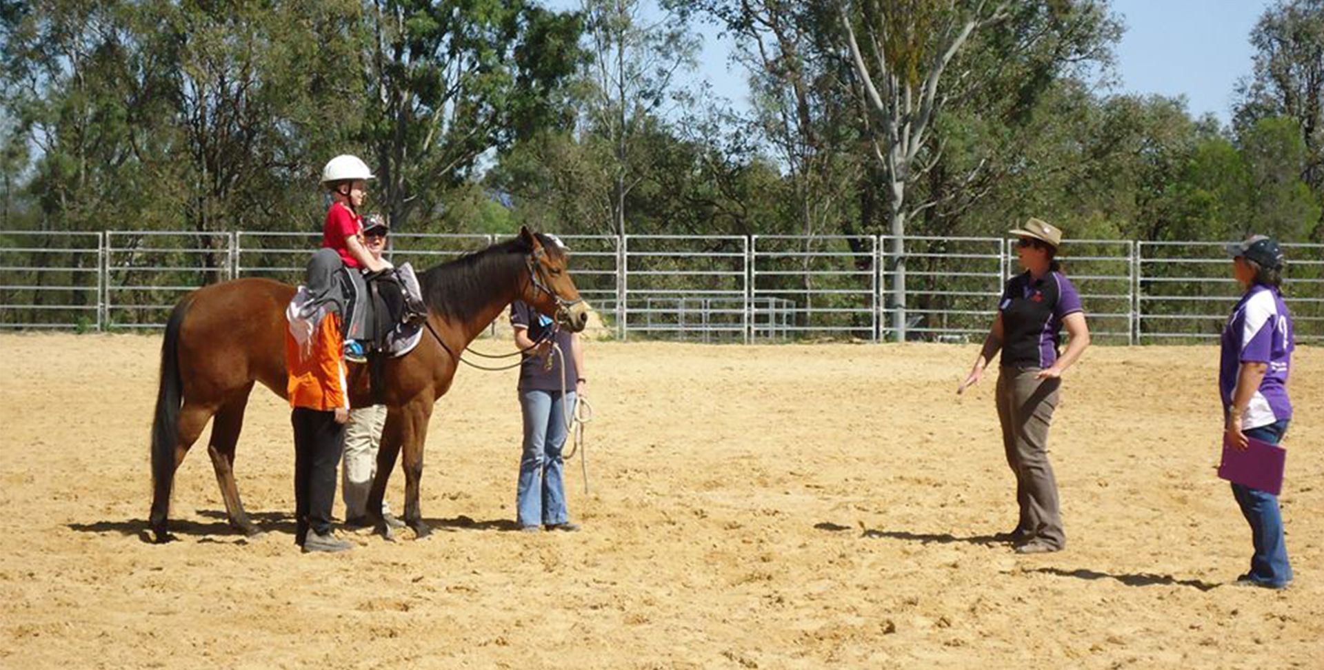 Child on a horse with two adults, one leading and one instructing in a sandy riding arena.