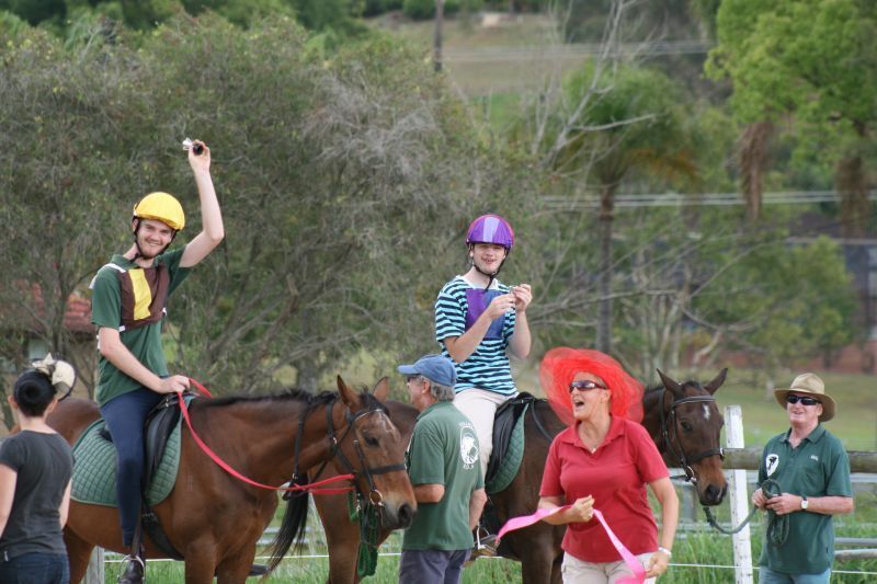 People on horseback at an event. A rider celebrates with a raised arm, another checks a clipboard, others watch.