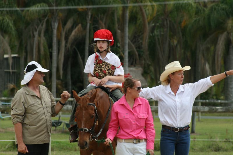 Girl on a horse with two assistants; one points forward, another holds the horse's lead.