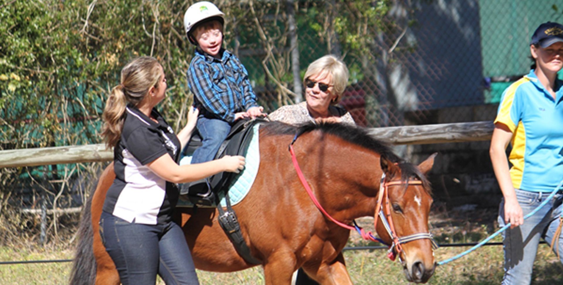 Boy with Down syndrome riding a brown horse, supported by two adults, outdoors.